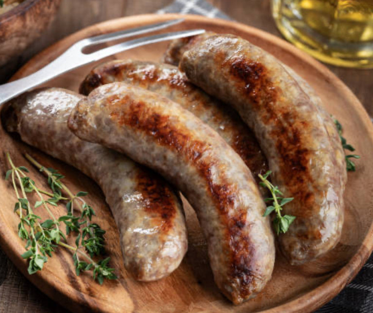 Four winterscattleranch Beef Brats are arranged on a wooden plate with fresh thyme. A metal fork and a blurred glass of oil appear in the background.