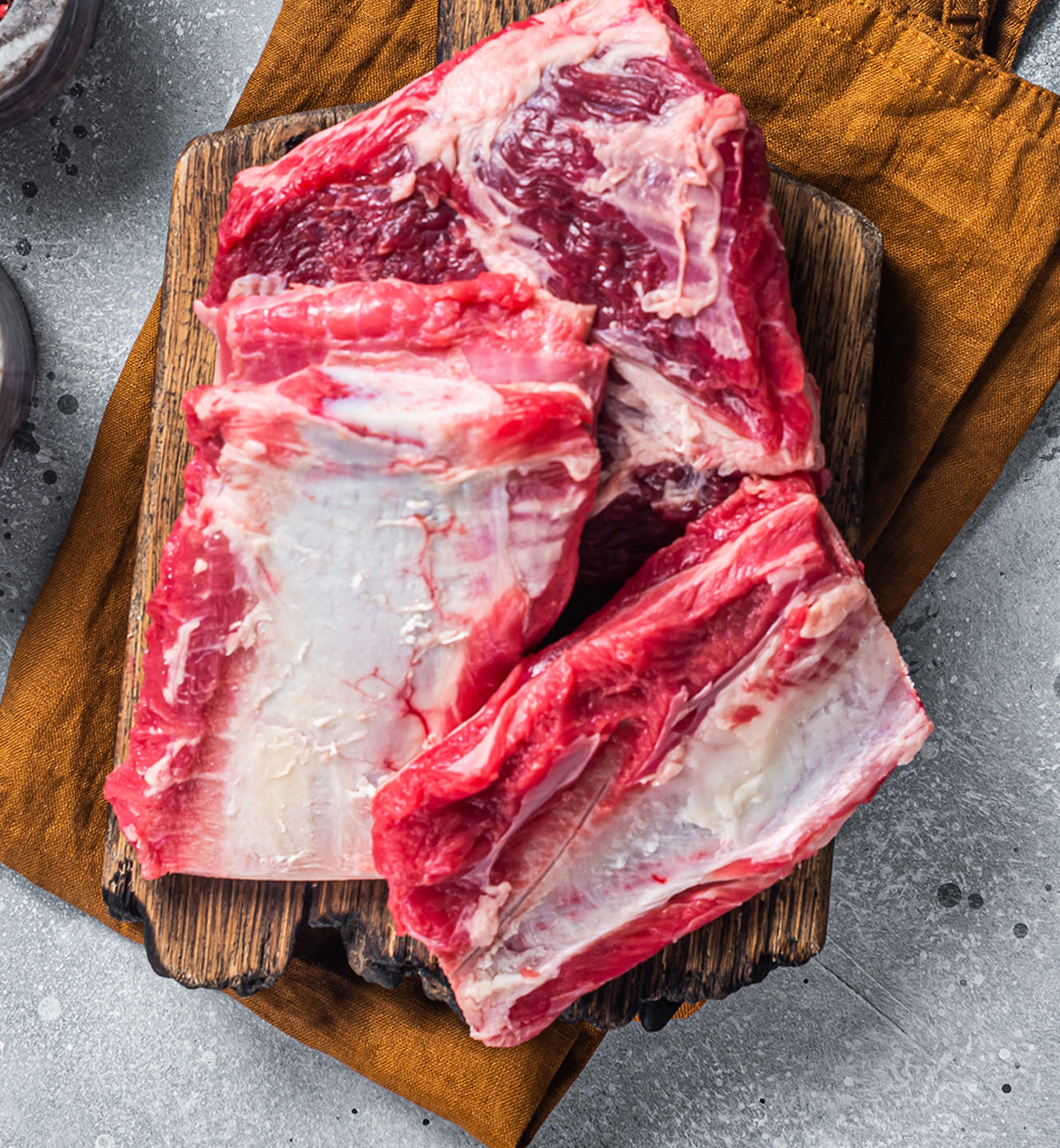 Winters Ranch, LLC Premium Organic Raised, Free-Range Grazing Beef Short Ribs are displayed on a wooden cutting board over a brown cloth and gray textured surface—ideal for braising or slow cooking.