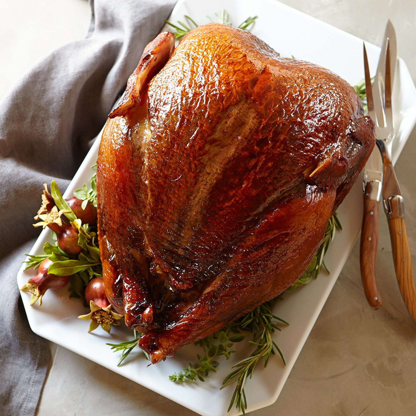 A golden-brown Winters Ranch, LLC Whole Fresh Turkey Deposit rests on a white platter, garnished with fresh herbs and red fruits. A carving knife and fork lie beside it on a beige surface next to a folded gray napkin.