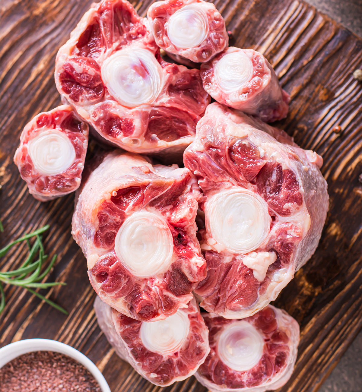 Winters Ranch, LLC Oxtail deposits feature marbled red meat and exposed bone, arranged on a wooden cutting board—perfect for stew. Fresh rosemary and a bowl of seasoning accompany this premium beef oxtail cut.