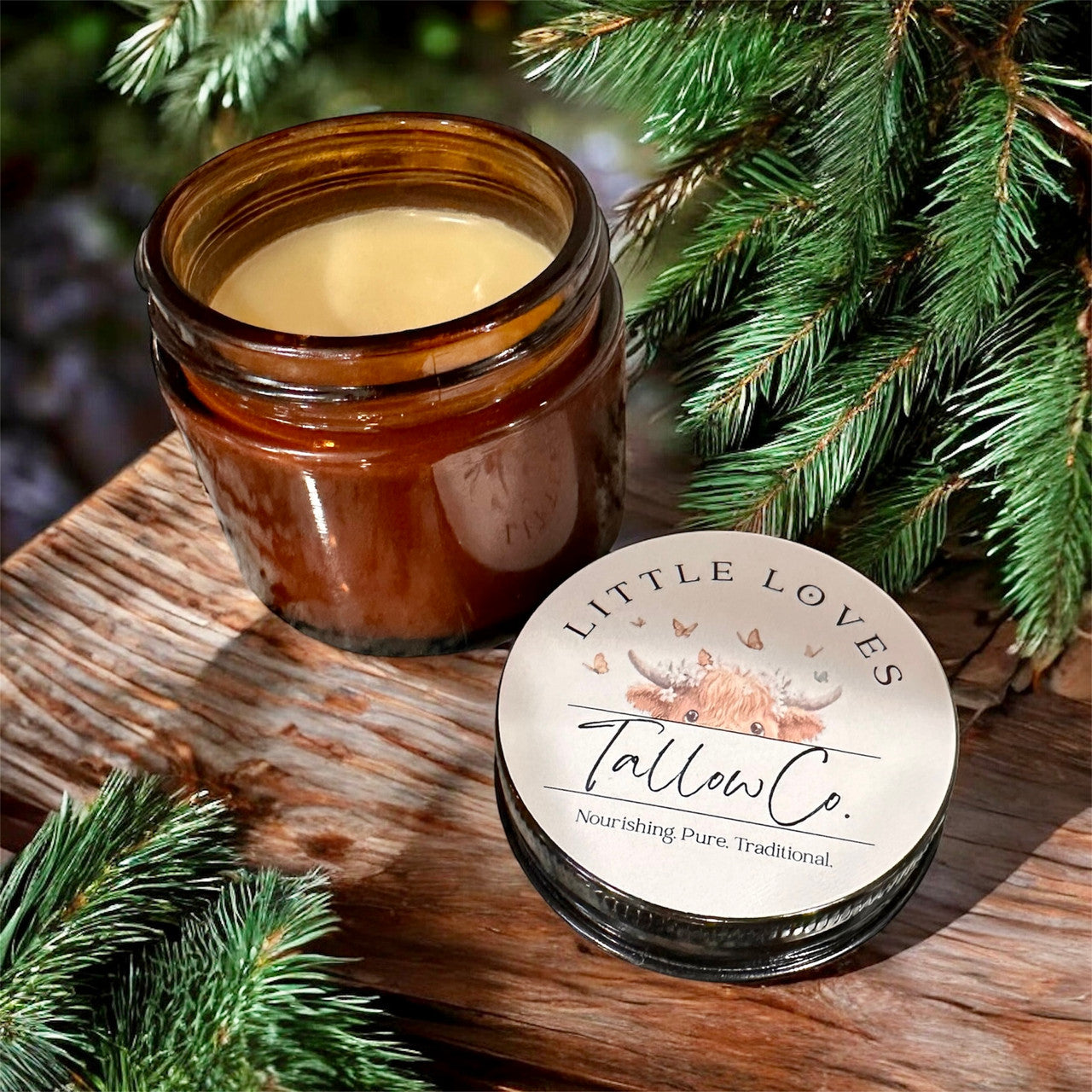 An open brown glass jar of Tallow from Winters Ranch, LLC rests on a wooden surface amid pine branches. The lid, set beside the jar, highlights this pure and nourishing option for organic skincare.