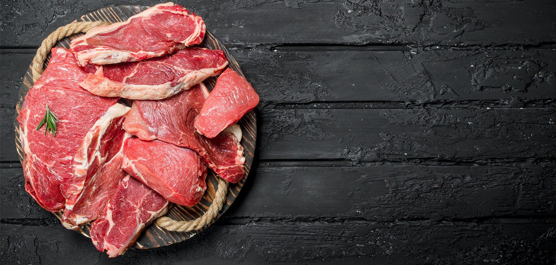 Several raw beef steaks are arranged on a round wooden platter with a sprig of rosemary, set on a dark rustic wooden surface.