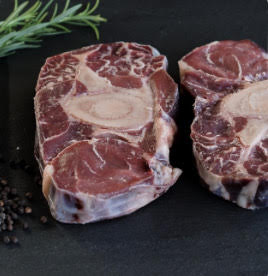 Two Soup Bones by winterscattleranch with visible bone and marbling, ideal for beef stock or soups, are placed on a dark surface with black peppercorns and a sprig of rosemary nearby.