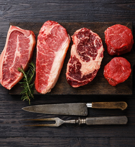 A wooden board with four raw steaks of different cuts, a sprig of rosemary, and vintage carving utensils on a dark wooden surface.
