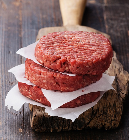 Three raw beef burger patties stacked with parchment paper between them, placed on a rustic wooden cutting board, with a wooden handle visible in the background.