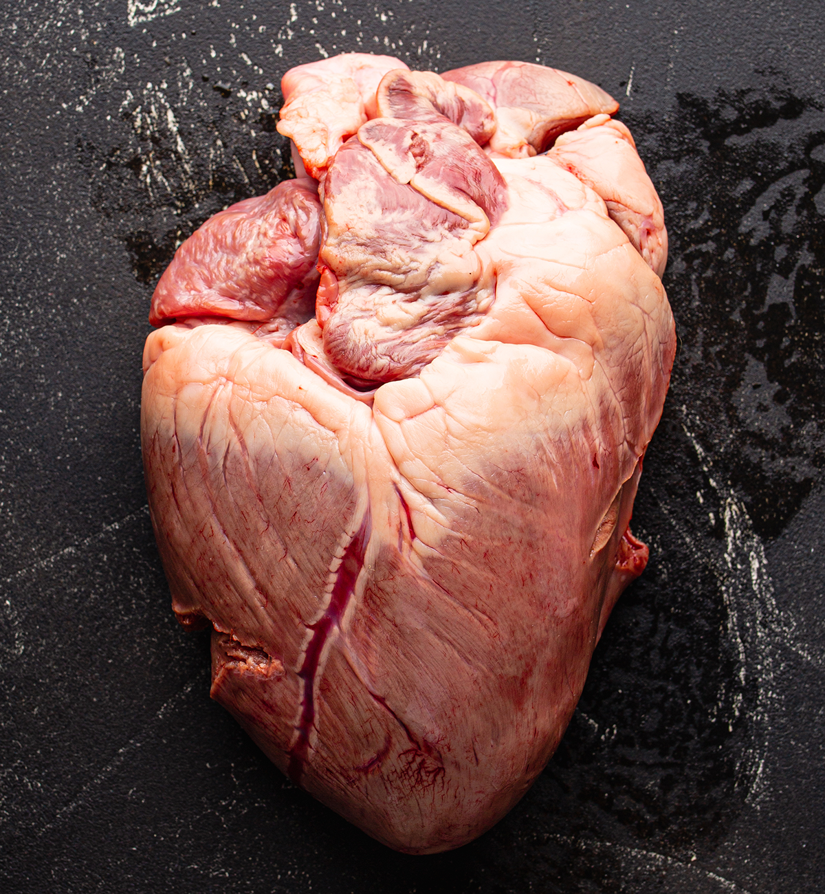 A large, raw Winters Cattle Ranch Beef Heart with visible veins and fat rests on a dark, textured surface, resembling a half pizza ready to be sliced.