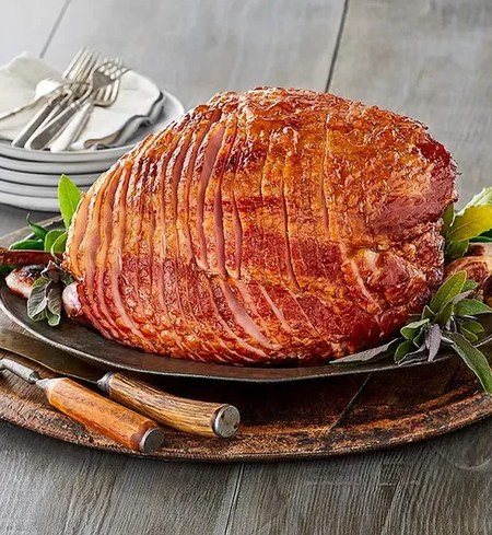 A large, glazed spiral-cut ham sits on a rustic platter, garnished with fresh herbs. Plates, forks, and knives are arranged nearby on a wooden table.