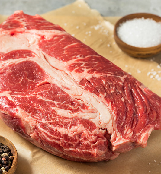 A raw Chuck Roast deposit from winterscattleranch rests on brown parchment, its rich red color and marbling ideal for a tasty pot roast or hearty beef stew, with bowls of coarse salt and peppercorns nearby.