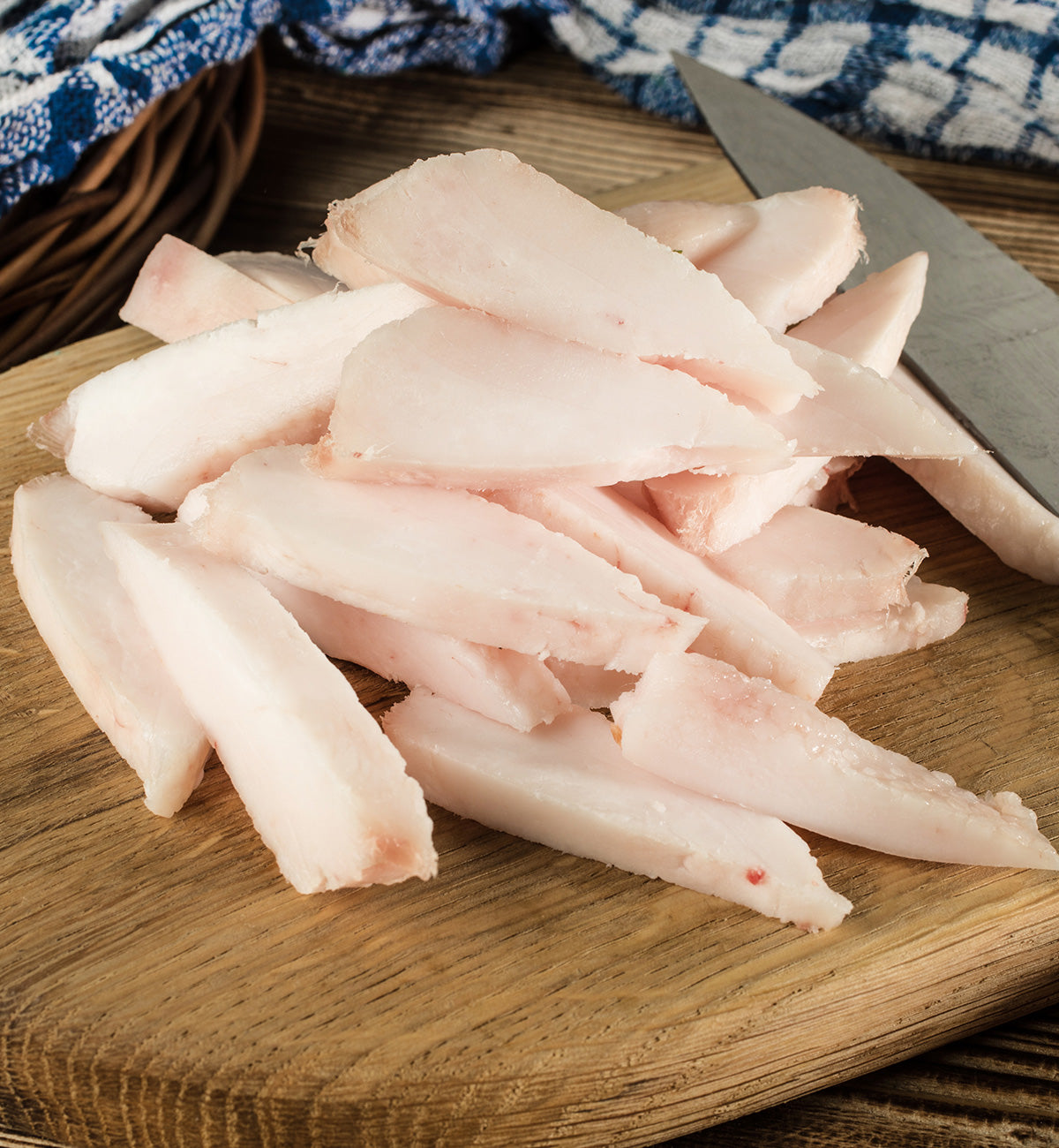 Slices of raw white chicken breast on a cutting board with a knife, blue and white checkered cloth in the background, and winterscattleranch Pork Fat in a bowl nearby.