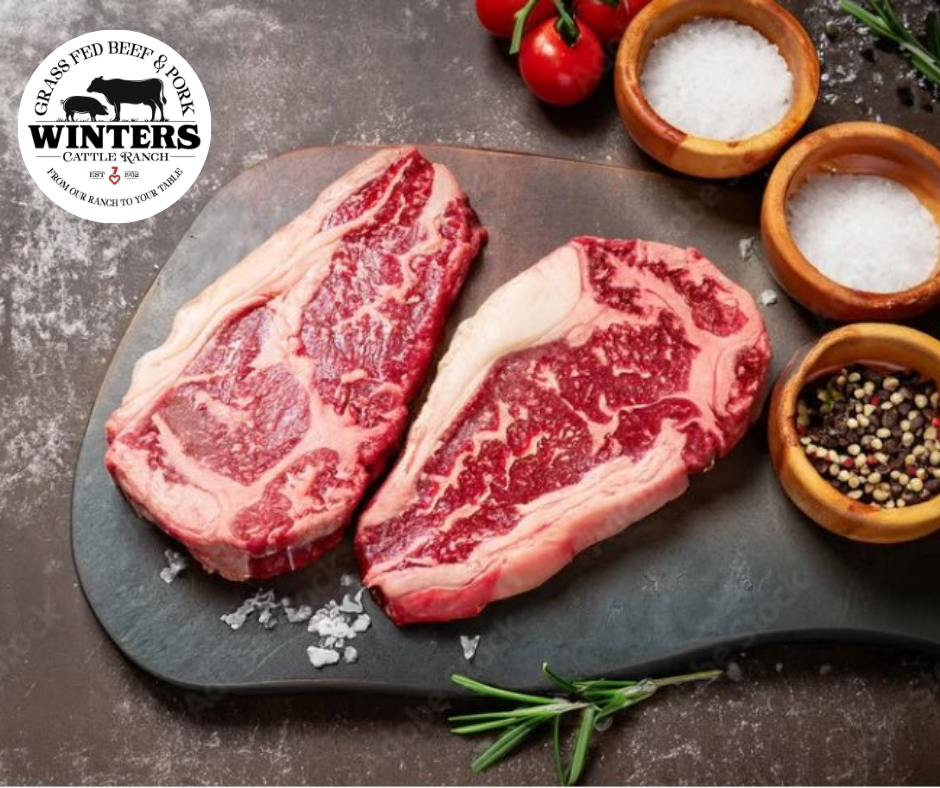 Two raw, marbled Ribeye steaks from winterscattleranch rest on a dark cutting board, surrounded by bowls of salt, pepper, herbs, fresh tomatoes, and sprigs of rosemary. A winterscattleranch logo appears in the top left corner.
