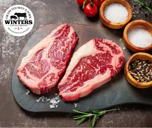 Two raw, marbled Ribeye steaks from winterscattleranch rest on a dark cutting board, surrounded by bowls of salt, pepper, herbs, fresh tomatoes, and sprigs of rosemary. A winterscattleranch logo appears in the top left corner.