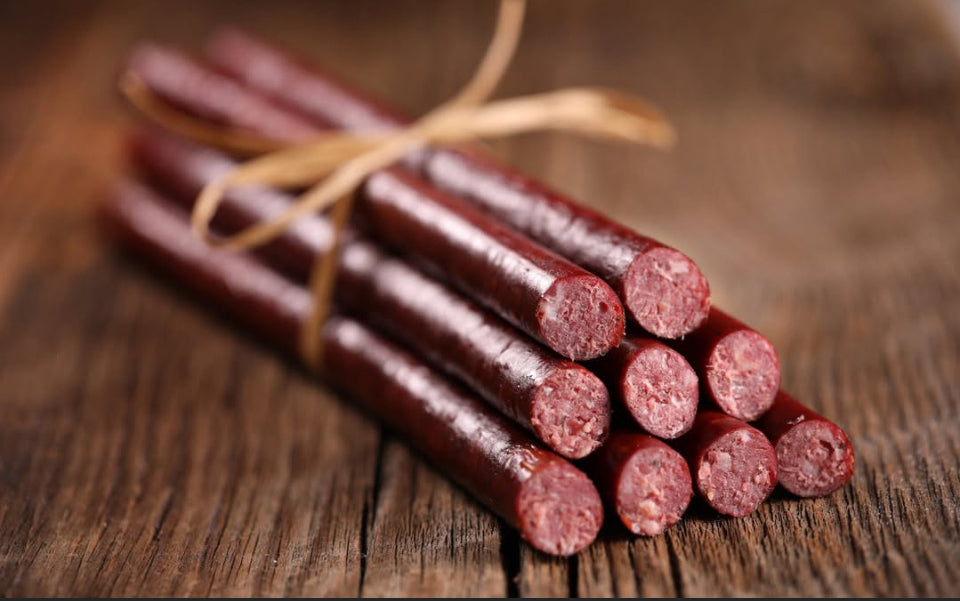 A six-pack of winterscattleranch Beef sticks, crafted from grass-fed Black Angus beef, is tied with twine and arranged on a rustic wooden surface, with the ends of the sausages facing forward to showcase the meat’s texture.