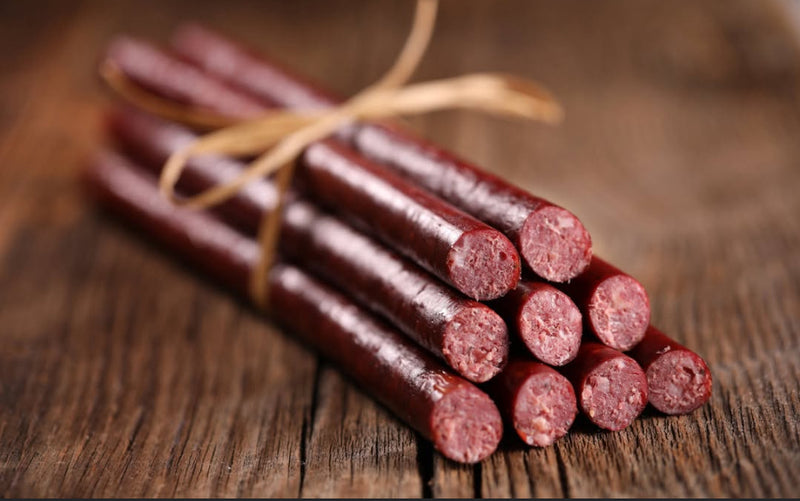 A six-pack of winterscattleranch Beef sticks, crafted from grass-fed Black Angus beef, is tied with twine and arranged on a rustic wooden surface, with the ends of the sausages facing forward to showcase the meat’s texture.