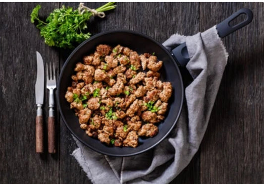 A black frying pan filled with cooked winterscattleranch Breakfast sausage 10 lb Subscription is garnished with parsley on a gray cloth, with fork and knife beside it. Fresh parsley rests above the pan on a dark wooden table.