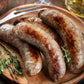 Six savory Beef Brats from winterscattleranch are served on a wooden plate, garnished with fresh thyme, beside a metal fork and a glass of yellow drink in the background.
