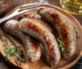 Six savory Beef Brats from winterscattleranch are served on a wooden plate, garnished with fresh thyme, beside a metal fork and a glass of yellow drink in the background.