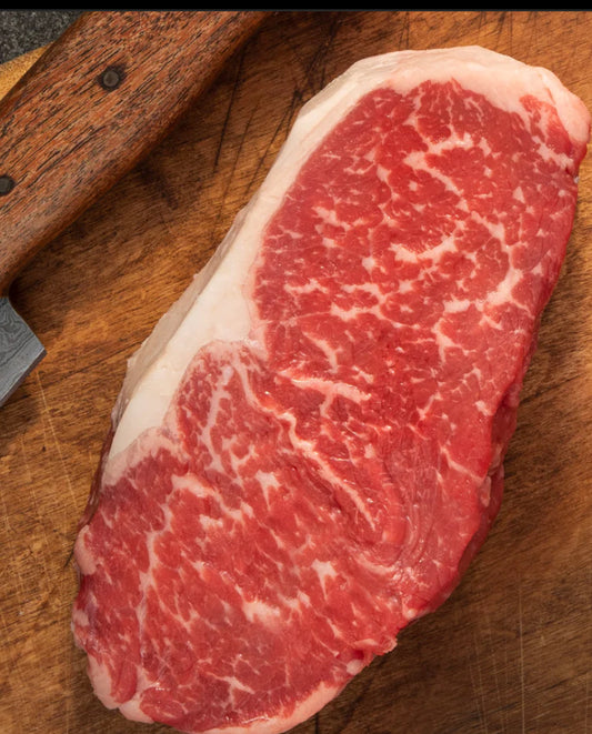 A winterscattleranch New York strip steak, richly marbled and grass-fed, sits on a wooden cutting board beside a wooden-handled knife.