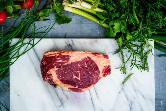 A winterscattleranch Ribeye steak rests on a white marble slab, surrounded by fresh chives, parsley, rosemary, green onions, and radishes.