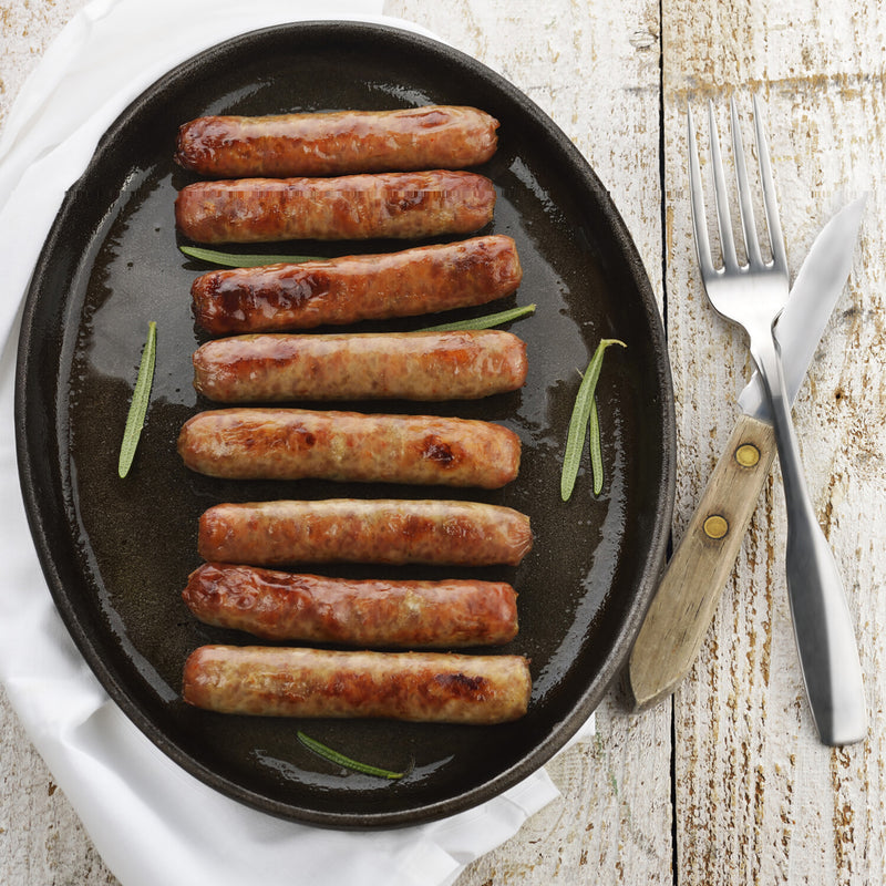 Eight Winters Cattle Ranch Breakfast Sausage Links, made from Berkshire pork, are arranged on a black oval plate with rosemary sprigs atop a white cloth. A fork and knife sit beside the plate on a rustic wooden table.