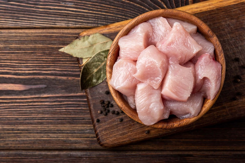A wooden bowl of raw, cubed Chicken Organically raised from Winters Ranch, LLC is on a cutting board with bay leaves and black peppercorns nearby, set against a rustic wooden background.