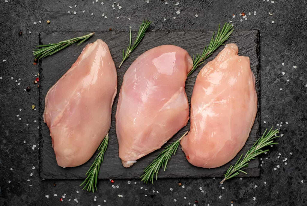 Three raw Chicken Organically raised breasts from Winters Ranch, LLC are displayed on a black slate board with fresh rosemary, set on a dark surface sprinkled with salt and peppercorns. Price is per pound.