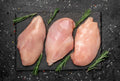 Three raw Chicken Organically raised breasts from Winters Ranch, LLC are displayed on a black slate board with fresh rosemary, set on a dark surface sprinkled with salt and peppercorns. Price is per pound.