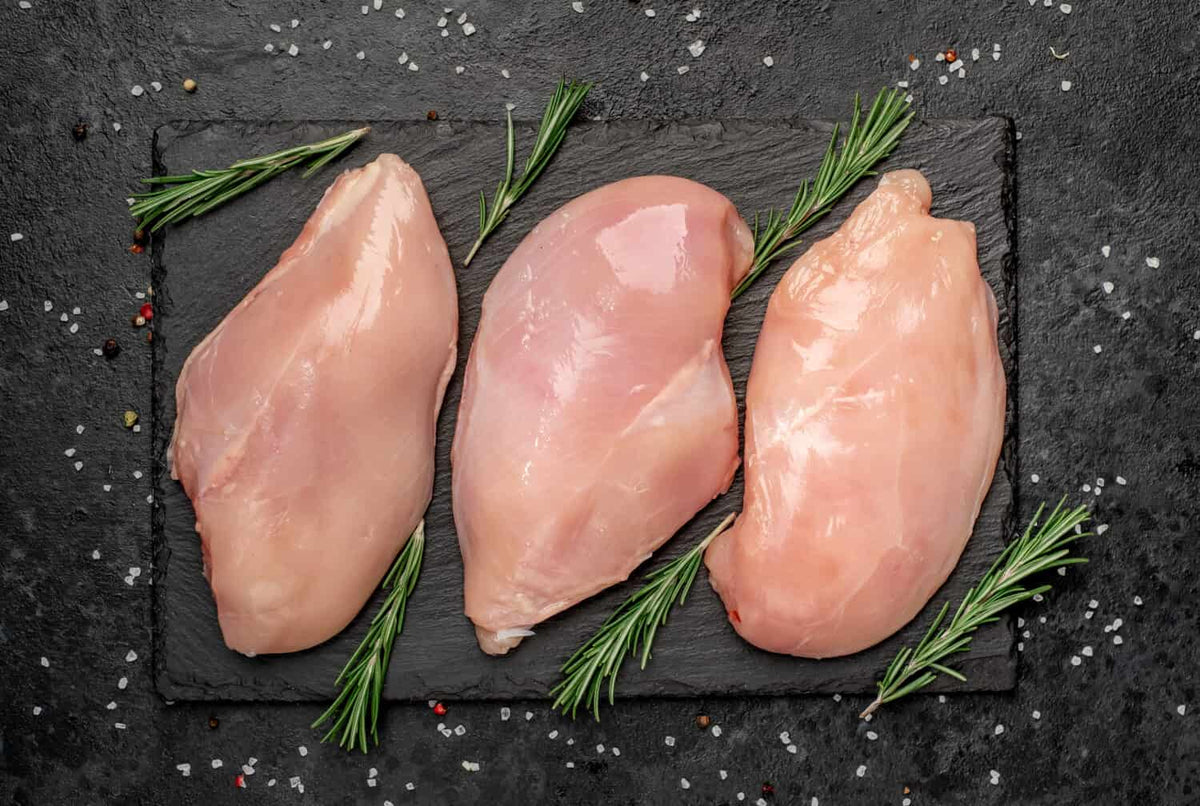 Three raw Chicken Organically raised breasts from Winters Ranch, LLC are displayed on a black slate board with fresh rosemary, set on a dark surface sprinkled with salt and peppercorns. Price is per pound.