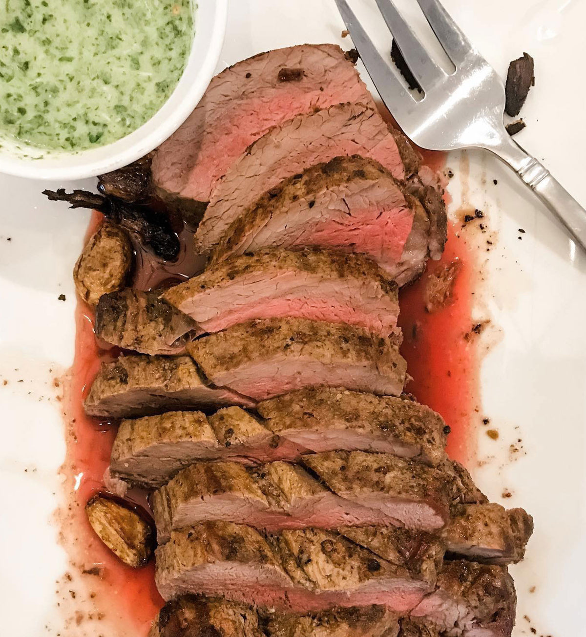 Winters Cattle Ranch Beef Tenderloin Roast deposits, sliced medium-rare, are served on a white plate with roasted garlic, green herb sauce, and a fork. Juices from this premium-grade beef enhance the presentation.