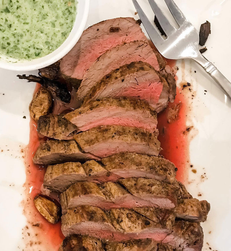 Winters Cattle Ranch Beef Tenderloin Roast deposits, sliced medium-rare, are served on a white plate with roasted garlic, green herb sauce, and a fork. Juices from this premium-grade beef enhance the presentation.