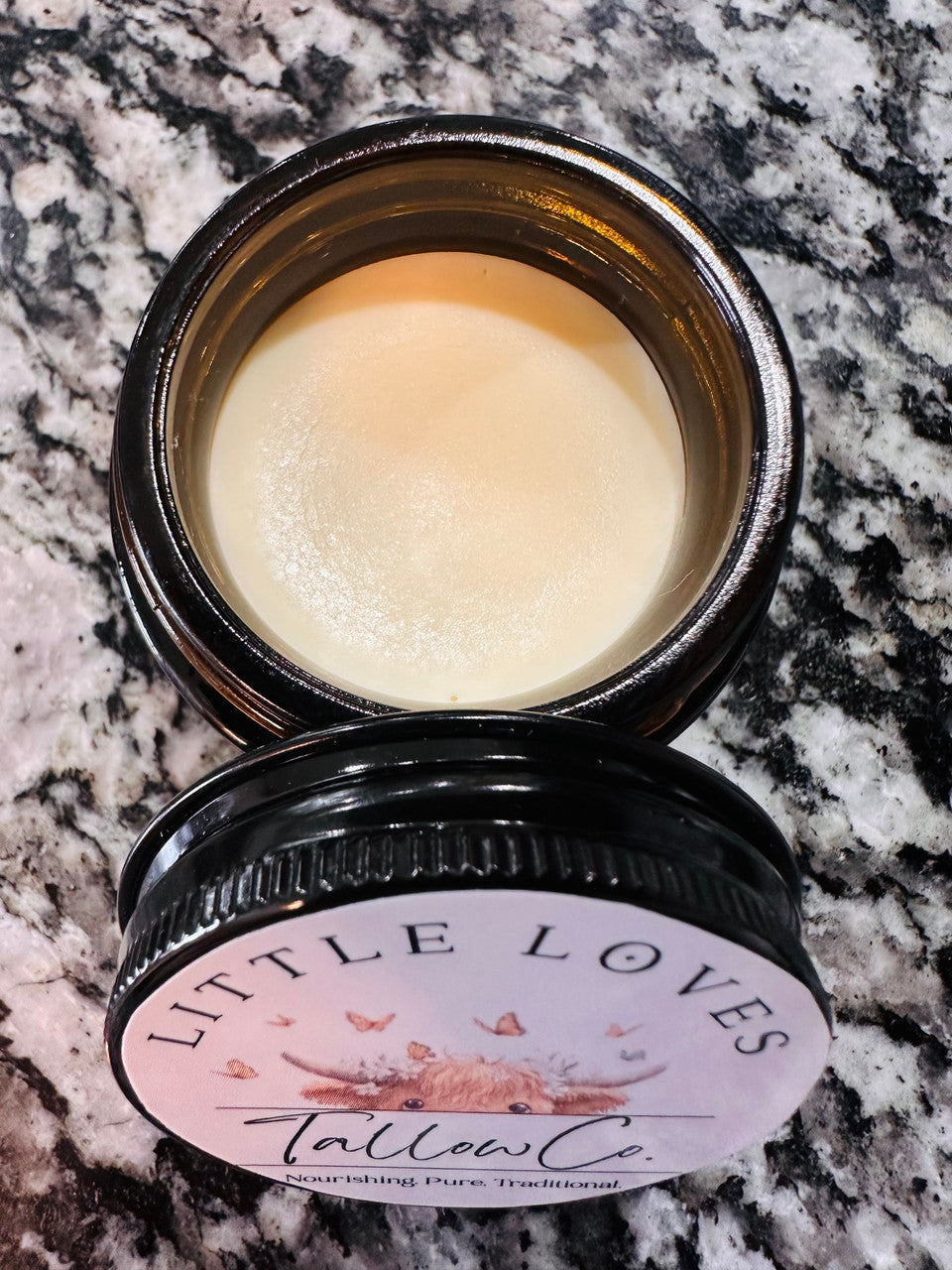 An open dark glass jar labeled Winters Ranch, LLC Tallow sits on a speckled marble countertop, filled with smooth balm. The label features butterfly and flower illustrations, making it perfect for natural skincare routines.