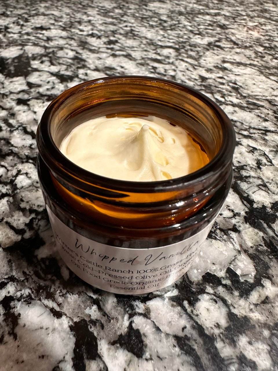 An open amber glass jar of Winters Ranch, LLC Tallow whipped balm rests on a marbled granite countertop. A white label identifies it as an organic skincare product.