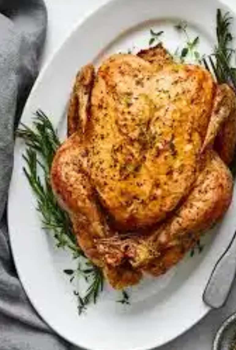 A whole roasted Winters Ranch, LLC Chicken Organically raised (price is per pound) with golden, crispy skin sits on a white platter, garnished with fresh rosemary and thyme sprigs.