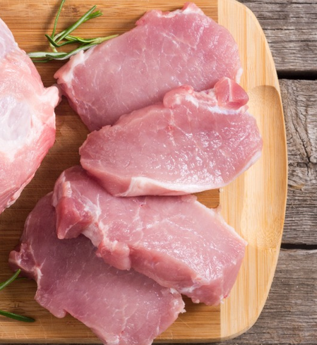 Four juicy Organic Raised Pork Chops (4 pack), bone-in, from winterscattleranch are arranged on a wooden cutting board with sprigs of fresh rosemary, placed on a rustic wooden surface.
