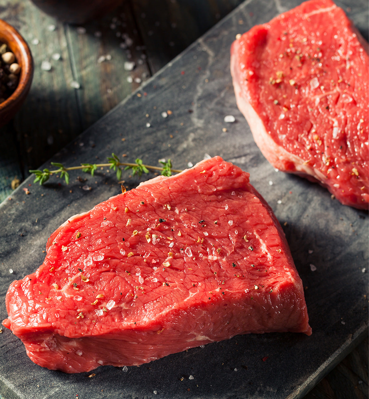 Winterscattleranch Sirloin steaks, seasoned with salt and pepper, rest on a dark stone surface. Fresh thyme sits beside the sustainable meat, with a bowl of peppercorns in the upper left corner.