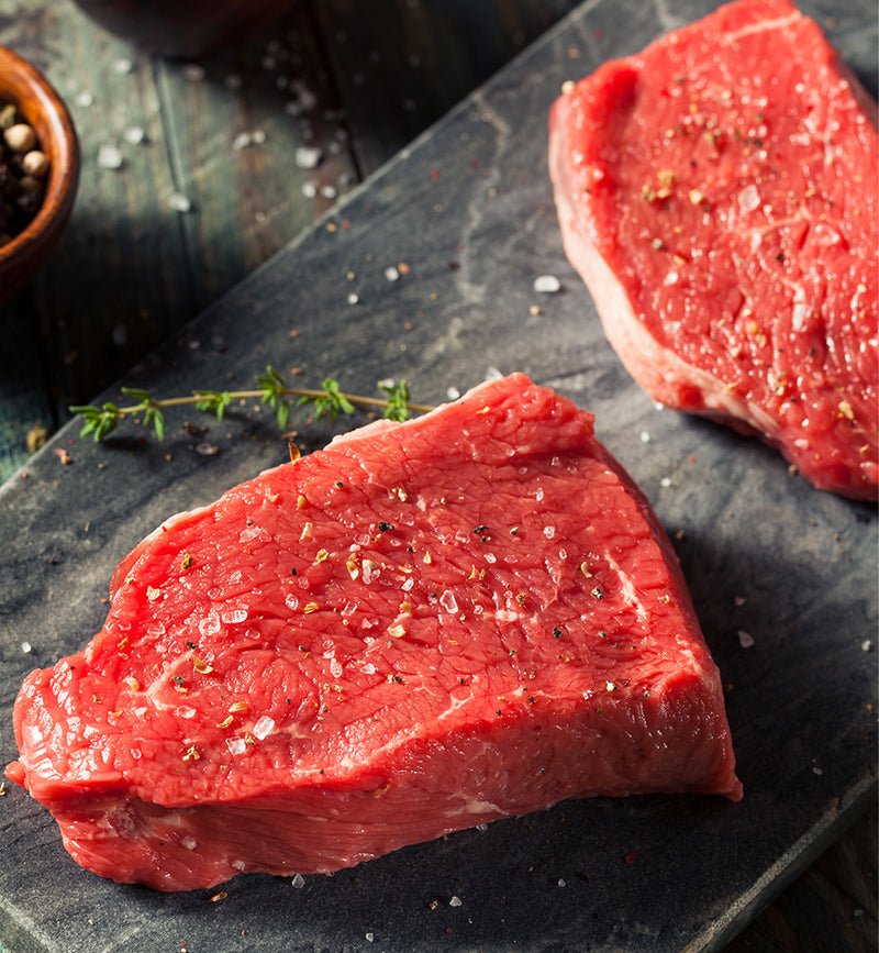 Winterscattleranch Sirloin steaks, seasoned with salt and pepper, rest on a dark stone surface. Fresh thyme sits beside the sustainable meat, with a bowl of peppercorns in the upper left corner.