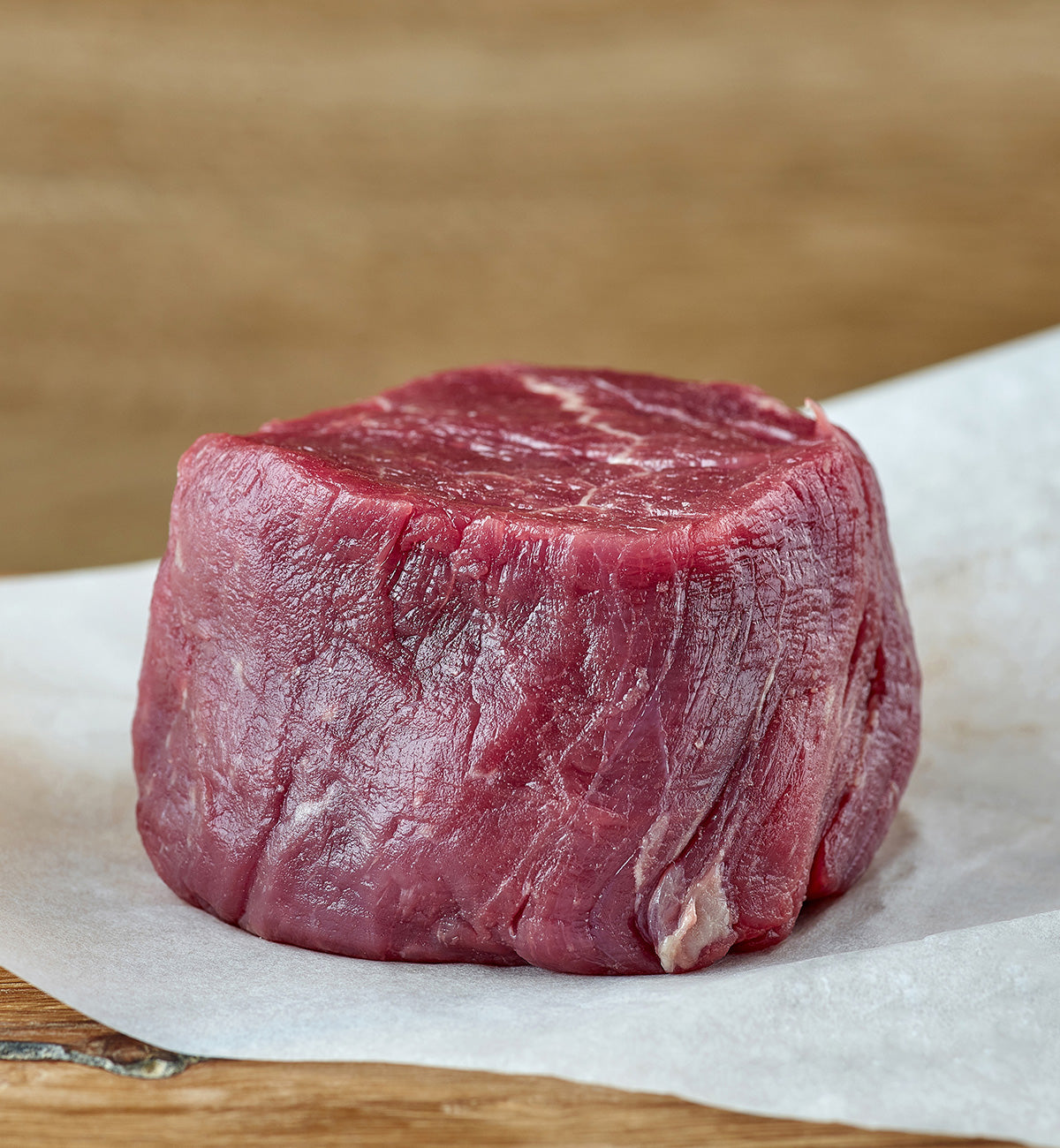 A winterscattleranch Beef Filet, marbled and fresh, sits on white parchment paper placed on a wooden surface, with a softly blurred brown background.